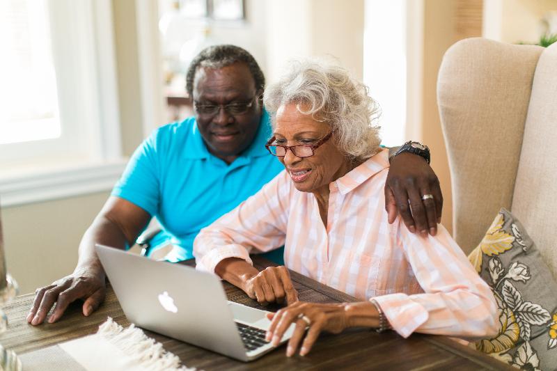 An elderly couple sitting at a table while using the laptop.