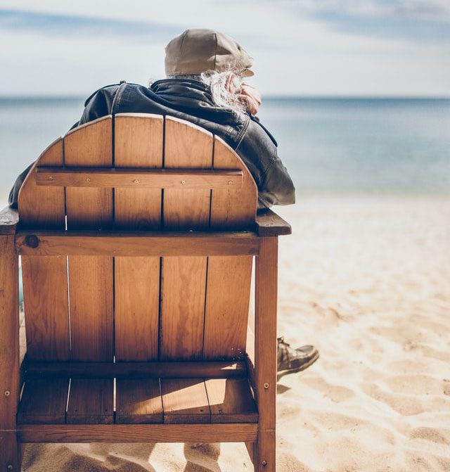An elderly man sits on a beach chair, facing the water.