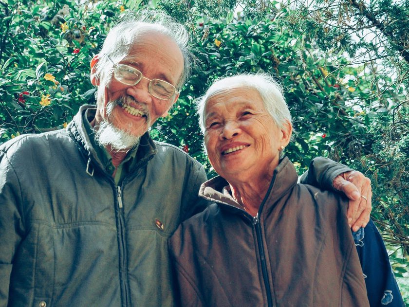 An elderly Asian couple in sweatshirts smiles for the camera.