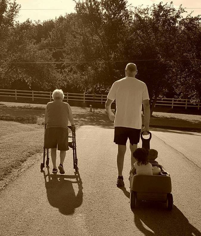 An elderly woman in a walker walks alongside an elderly man pulling a cart with two toddlers.