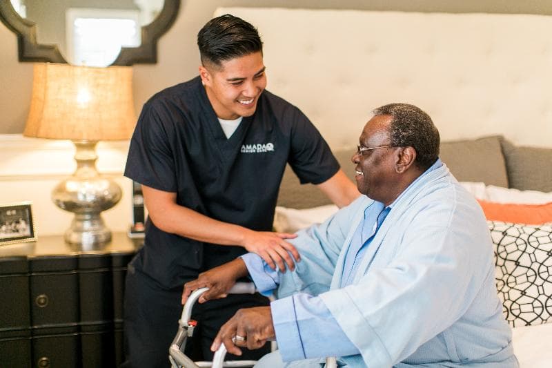 A young man in an Amada caregiver uniform assists an elderly man in standing from his bed to his walker.
