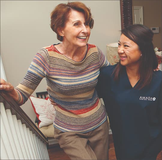 A cheerful elderly woman is helped up the stairs by an Amada caregiver.