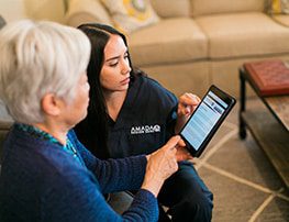 An elderly woman using a smart device is assisted by an Amada caregiver.