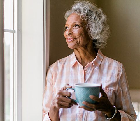 A happy-looking elderly woman holding a blue tea cup smiles outside the window.
