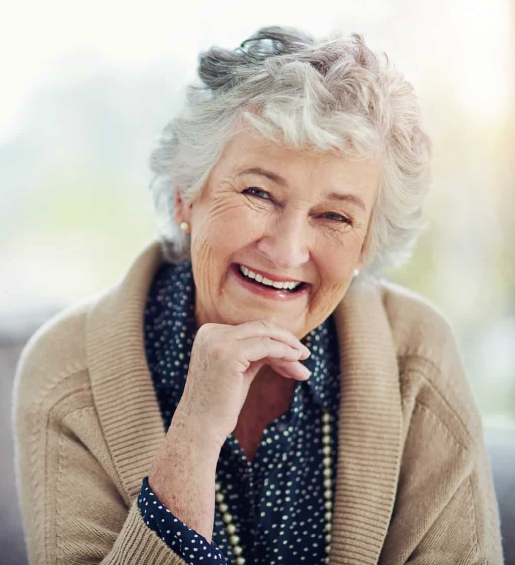 A cheerful elderly woman in a light brown coat smiles for the camera.