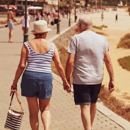 An elderly couple walks by the beach shore holding hands.