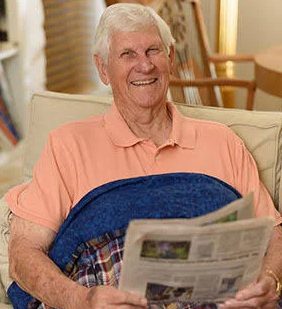An elderly man on a couch poses with a grin while reading a newspaper.