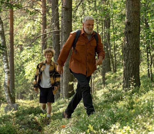 An elderly man walks ahead of a younger man in the woods.