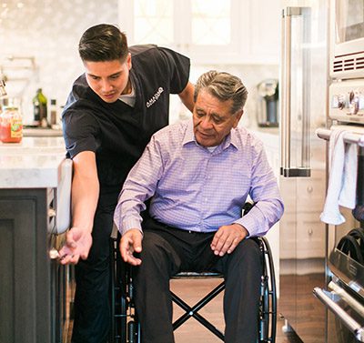 A young man dressed in an Amada caregiver uniform helps an elderly man in a wheelchair open a kitchen cabinet.