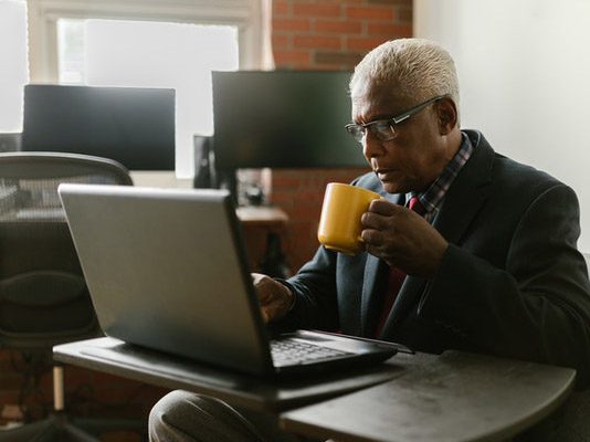 An elderly man in a suit sits at a desk with his laptop and a coffee mug.