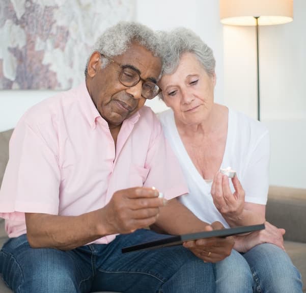 An elderly couple sits on a couch, holding candles and looking sadly at a picture frame.