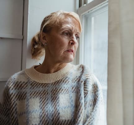 An elderly woman with a worried expression sits by the window.