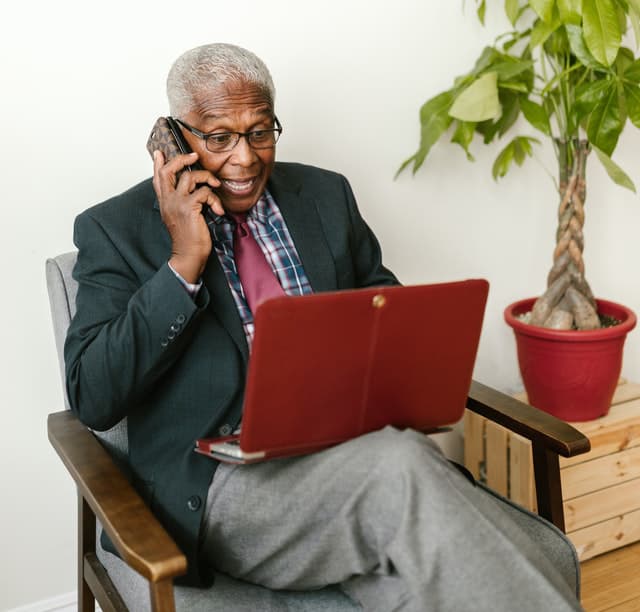 An elderly man in a suit, sitting on a chair, using his laptop and phone.