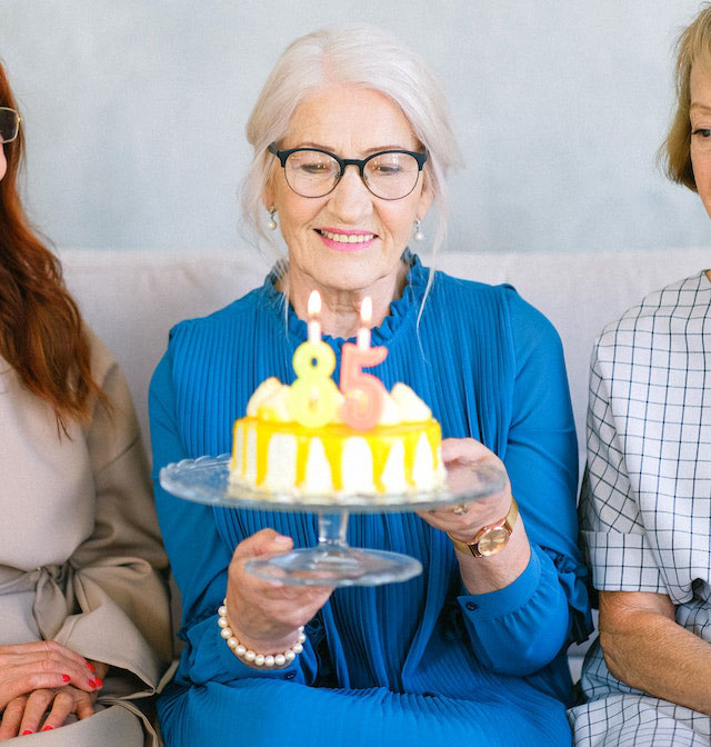 An elderly woman in a bright blue dress and eyeglasses smiles as she holds a cake with an eighty-five candle topper.