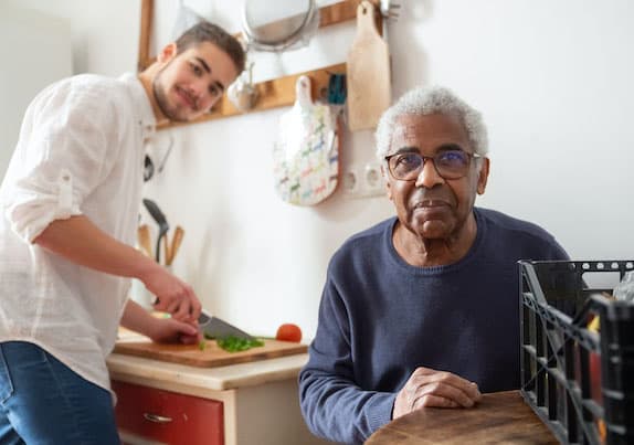 A younger man preparing food for an elderly man while looking at the camera.