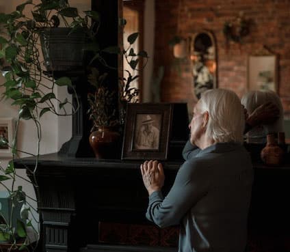 A sad elderly woman looks at a picture frame of a man on the table.