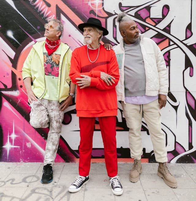 Three elderly males posing stylishly in front of a vibrant wall backdrop.