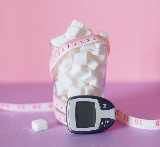 A clear glass with sugar cubes in it, wrapped in a tape measure and a glucose meter beside it.