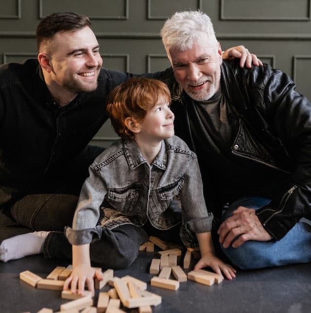 A child sitting on the floor with his grandfather and dad, laughing, playing happily with wooden block tiles.