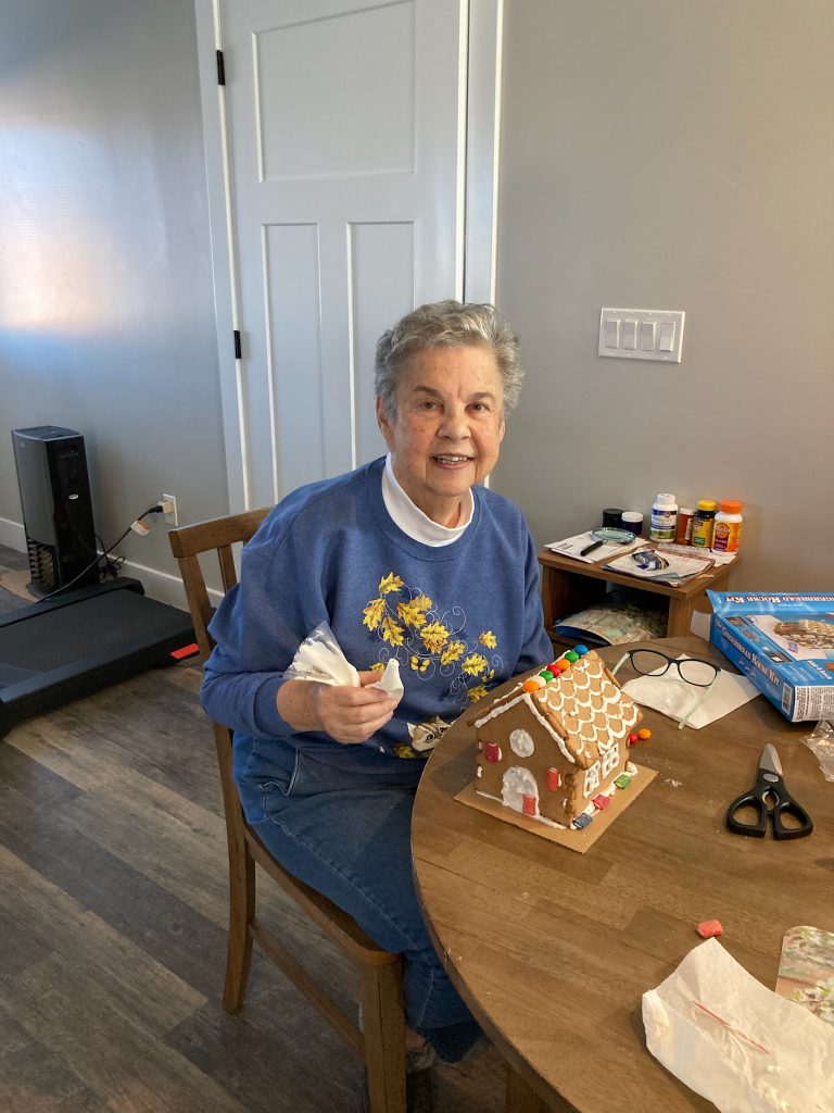 An elderly woman works on a gingerbread house at a table.