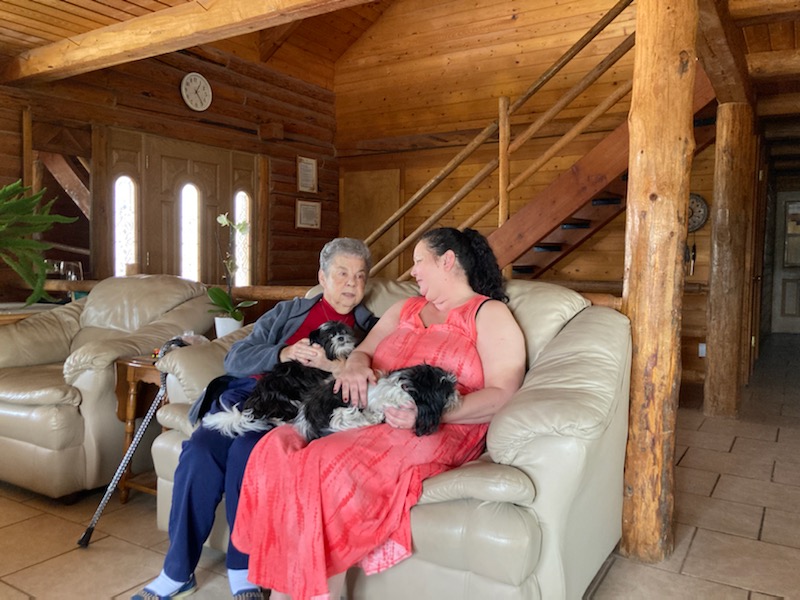 An elderly mother and her daughter chat while sitting on a couch with their dogs on their laps.