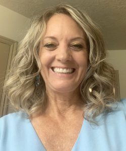 Margie4.5 copy A blonde-haired woman wearing a blue shirt smiling in front of a door.